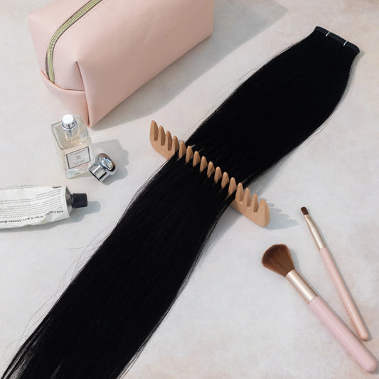 Black hair extensions on a light surface with cosmetic items including a pink bag, perfume, and brushes.